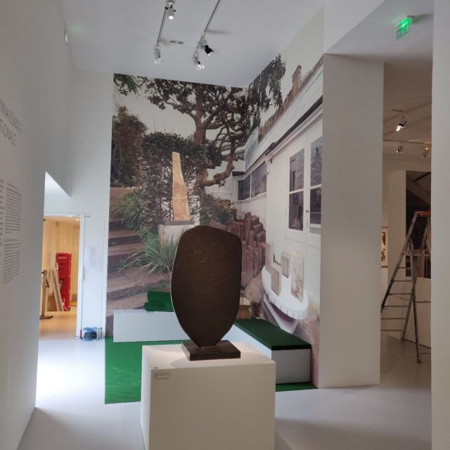 Salle d'exposition avec mural photographique de jardin et sculpture sur socle blanc